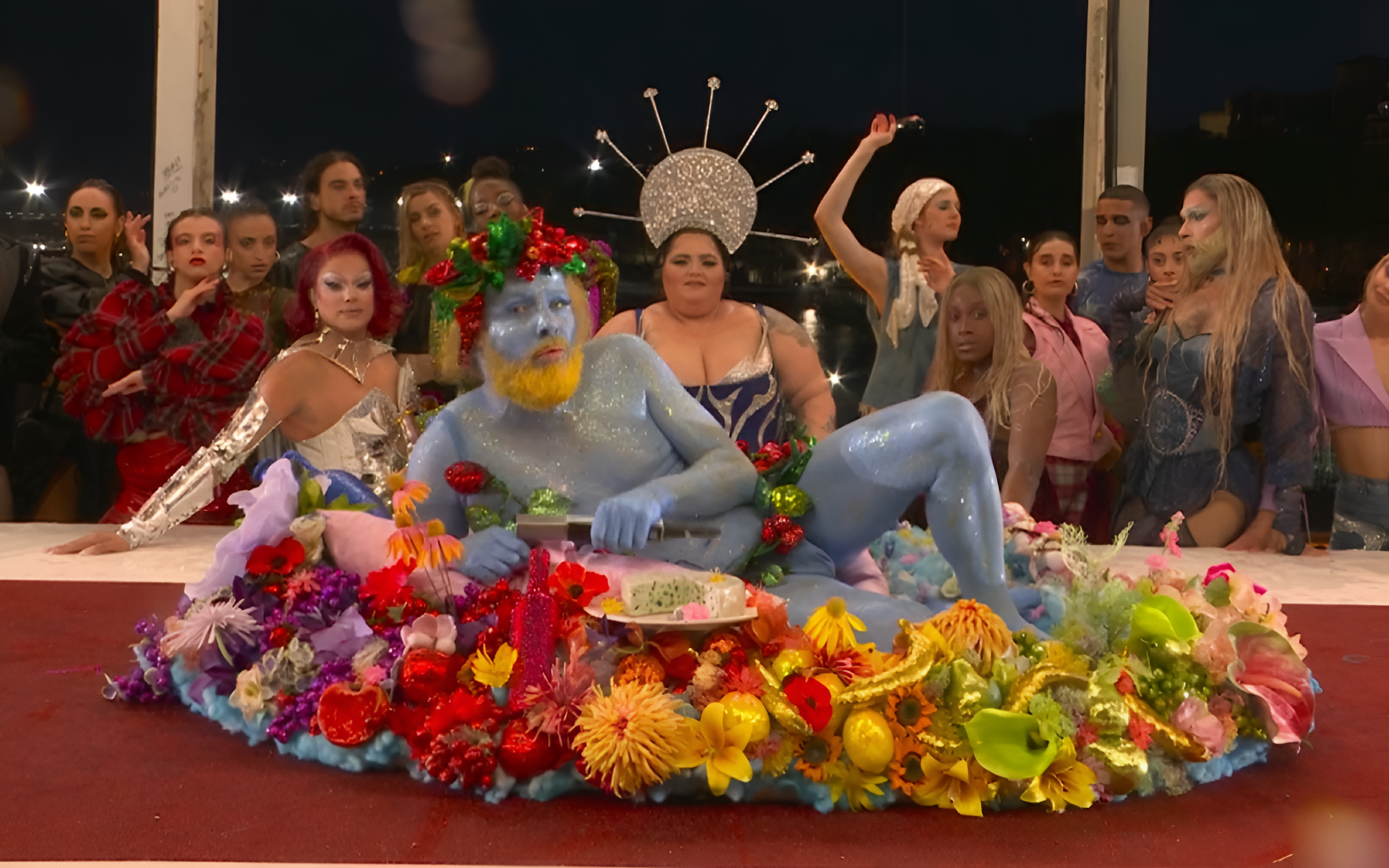 Un homme peint en bleu au front ceint d'une couronne de fleurs allongé sur une table couverte de fleurs ; derrière la table, une dizaine de personnes habillées de façon extravagante